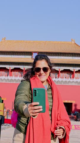 Happy woman wearing sunglasses and a scarf using her smartphone to take a self portrait with the historic Meridian Gate of the Forbidden City in Beijing, China, on a sunny day
