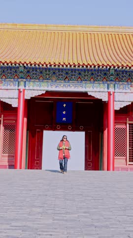 Young tourist woman exploring the ancient Forbidden City, walking towards the camera with a traditional chinese palace in the background during a sunny day in Beijing, China, a famous landmark