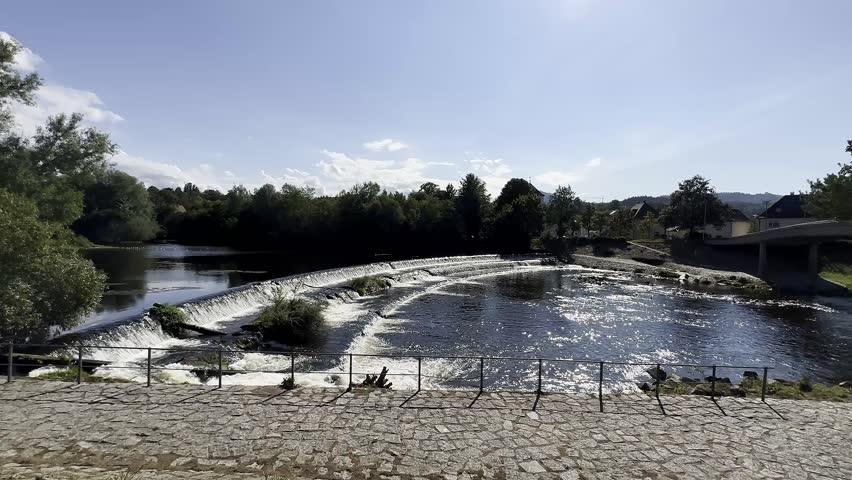 A 4K video clip showcasing a scenic river view with a waterfall. The camera pans across the landscape under bright daylight, highlighting natural beauty.