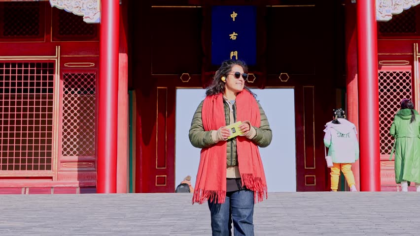 Female tourist with sunglasses and a red scarf smiling while listening to an audio guide in front of a traditional chinese building at the Forbidden City, a famous landmark in Beijing, China