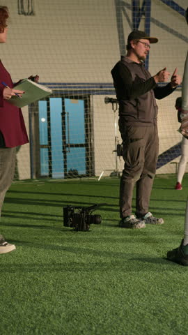 Full vertical shot of Caucasian female producer and cameraman directing crew and actors while checking script and storyboard, filming sports show for television, standing on green turf at stadium