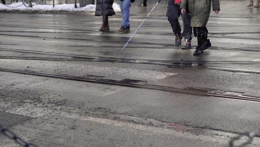People crossing road at crosswalk, person using cane navigating city.