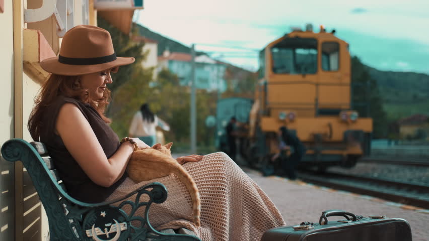 Woman sits calmly at train station with cat on lap. She wears hat, looks relaxed. Train waits in background. Suitcase rests beside her. Scene suggests travel or waiting. Ideal for travel, relaxation
