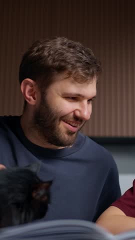 Young man with beard and short hair wearing a dark shirt sitting indoors and interacting with a black cat in a cozy environment with a brown wooden wall background