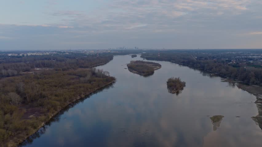 Serene aerial view of meandering Vistula River with forested islands near Warsaw. Calm water reflects sky with distant city skyline visible
