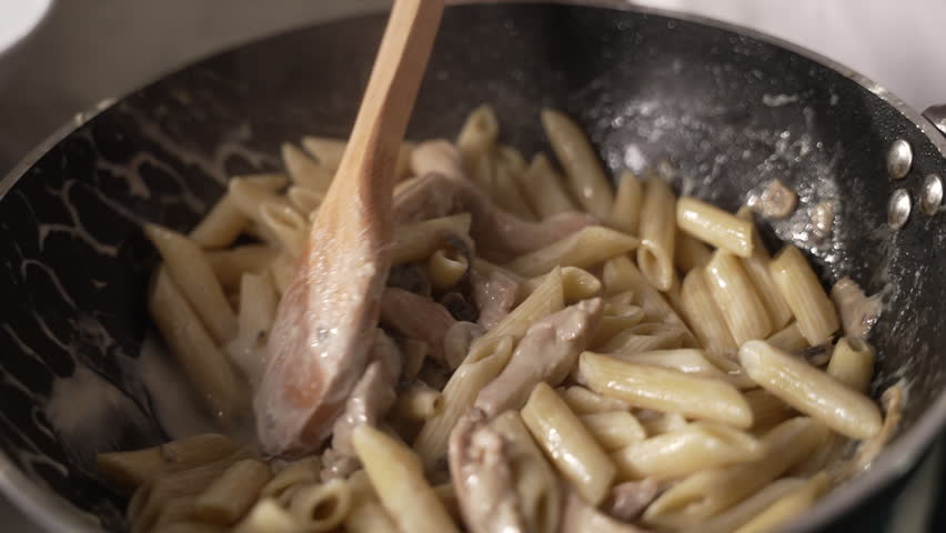 Wooden spoon stirring creamy penne pasta with chicken and mushrooms in a black frying pan, fa01
