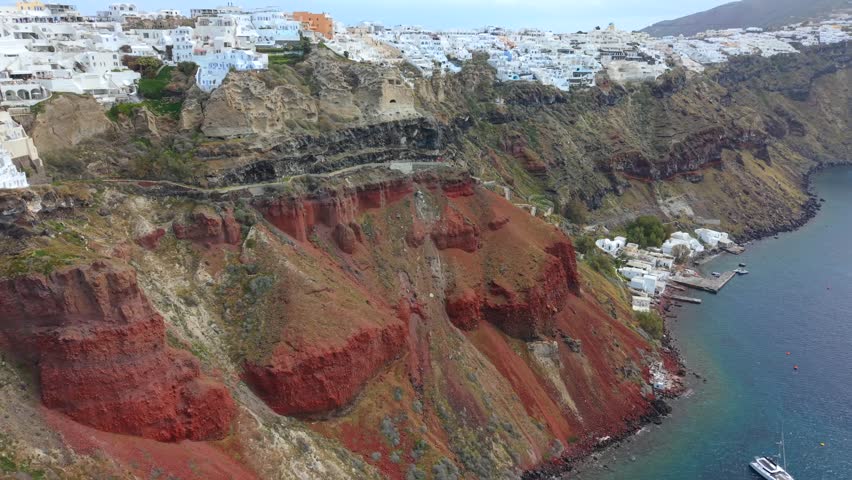 Aerial video looking down the vibrant red volcanic cliffs of Oia toward the small harbour of Armeni Bay, featuring catamarans anchored in the sapphire Aegean Sea.