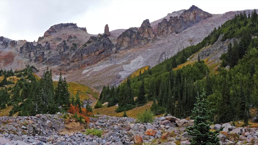 A wide-angle landscape shot of the Glacier Basin Trail, featuring a rugged, rocky riverbed in the foreground leading toward a dense coniferous forest. In the distance, the sharp, jagged ridges of the