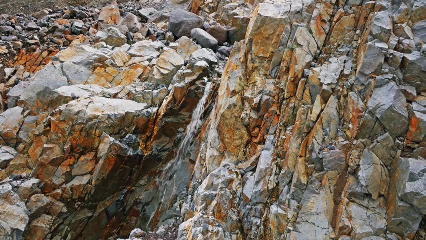 Detailed view of a fractured, weathered rock cliffside in Mount Rainier National Park, Washington State. The image features vibrant orange, rust, and grey geological textures, highlighting the rugged