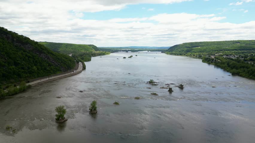 Distant high angle drone shot capturing a wide, calm river winding through lush green forested hills and valley landscapes on a clear day.