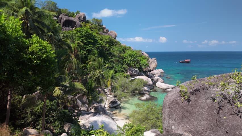 Lush palm trees and boulders frame a tranquil cove with turquoise water and a boat