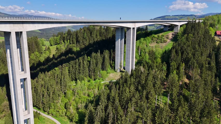 Aerial view of the Lavanttal Bridge spanning across a deep valley with dense pine forests and rolling green hills under a blue sky in Wolfsberg, Carinthia, Austria.
