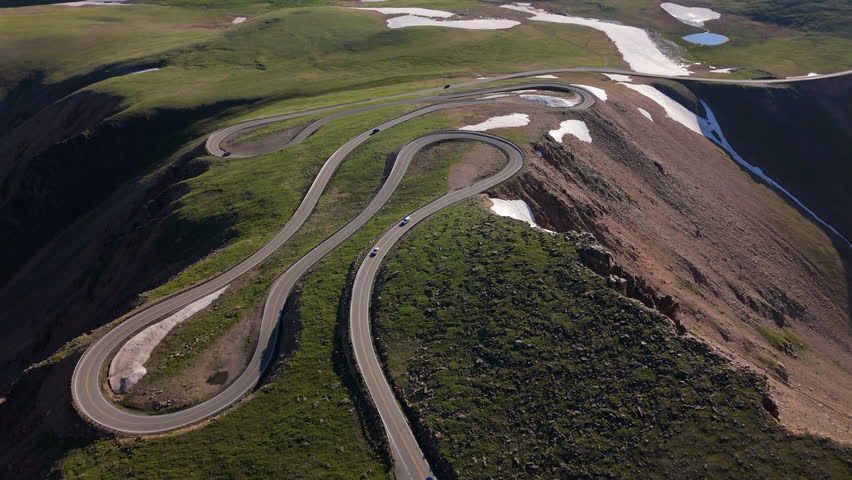 Aerial of Beartooth Highway in Montana USA reveals dramatic serpentine curves through alpine tundra. Drone captures winding mountain pass with scattered snow patches and green meadows on sunny day.