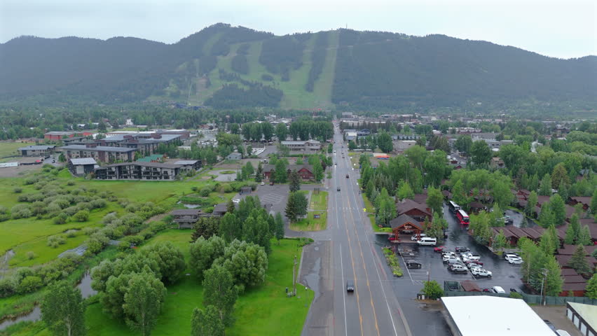 Aerial drone footage captures Jackson Hole Wyoming on a rainy day with lush green ski slopes visible on mountainside. Cars travel along the main road through town surrounded by hotels and pine trees.