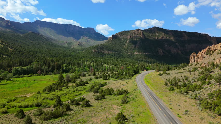 Aerial Montana USA view of winding mountain highway cutting through lush green valley with dramatic rocky cliffs and pine forests under bright blue summer sky with white clouds.
