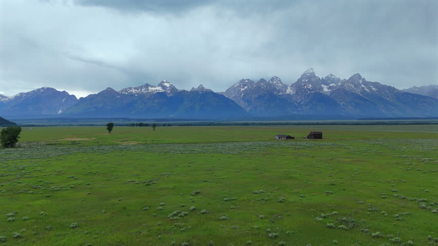 Aerial drone footage of Grand Teton National Park in Wyoming USA showcasing vast green prairie with rustic barn buildings and dramatic snow-capped mountain peaks under overcast skies.