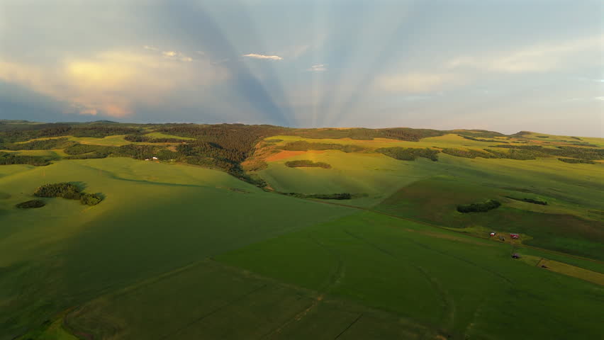 Aerial Montana USA landscape featuring vast rolling green hills and farmland under dramatic sky with crepuscular sunbeams radiating through clouds at golden hour. Pastoral countryside vista.