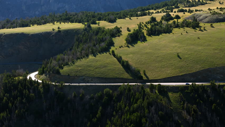 Aerial view of scenic mountain highway cutting through dramatic Montana landscape with vehicles traveling along winding road bordered by dense evergreen forests and expansive green alpine meadows.