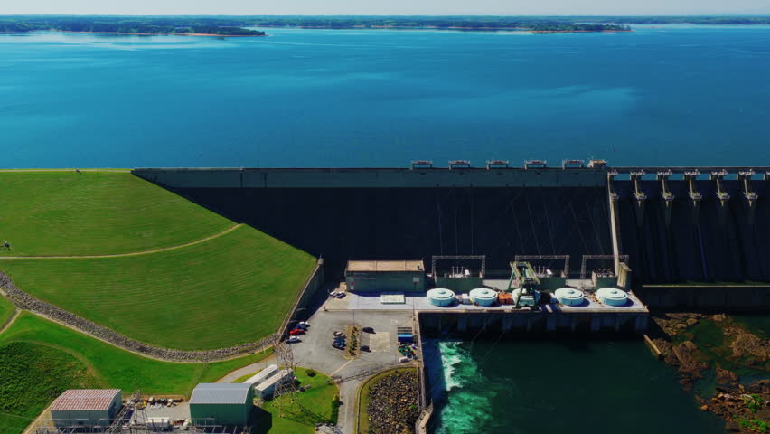 artwell Dam showing the grass-covered earthen embankment, concrete spillway, powerhouse with four turbine units, active water release, and Lake Hartwell reservoir with distant islands.