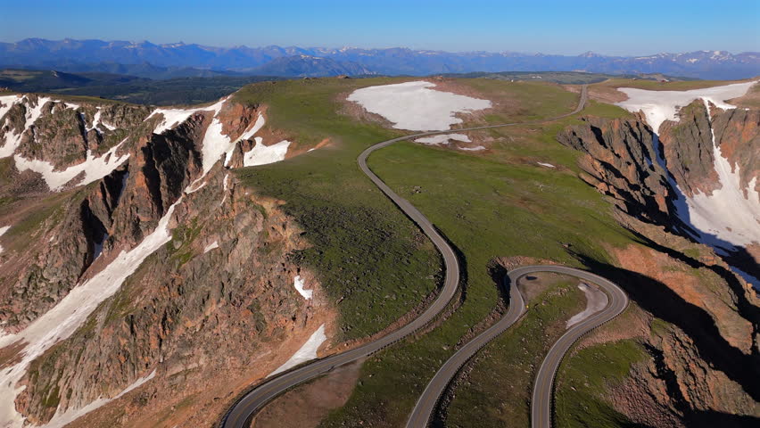 Aerial drone footage of Beartooth Highway winding through dramatic alpine landscape with snow patches dotting green meadows, rugged red rock cliffs, distant mountain ranges under clear blue summer.