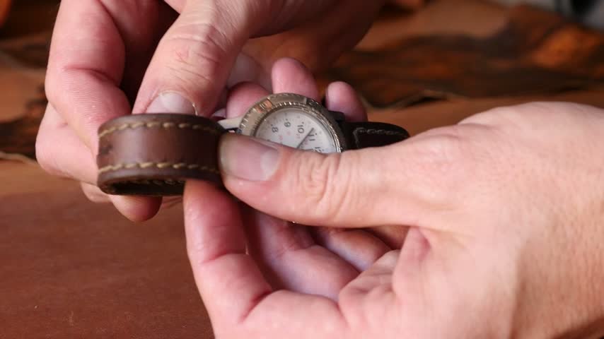 Macro shot of artisan hands examining a custom stitched brown leather watch band, checking the metal buckle, flexibility, and mounting it on a vintage wristwatch.