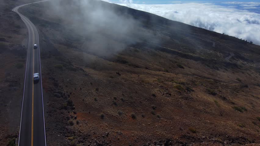 Drone pullback shot from the summit road of Haleakala volcano on Maui Hawaii with dramatic clouds rolling over the volcanic landscape above the clouds