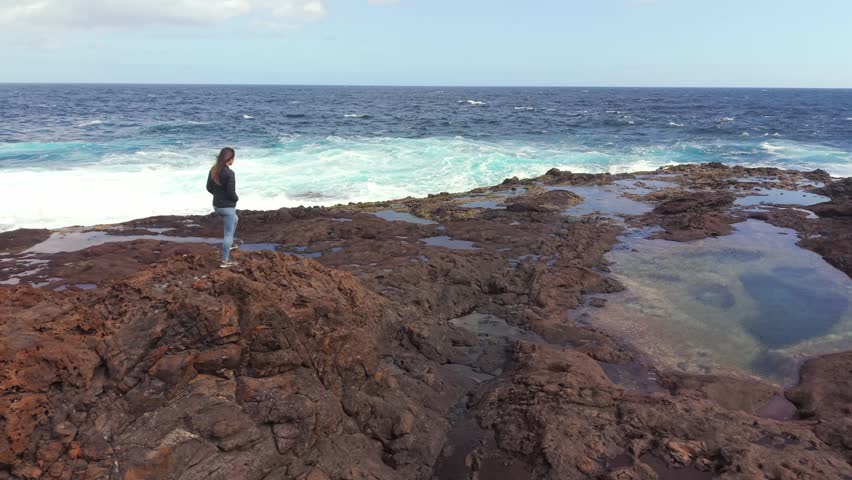Woman standing on a rocky volcanic shoreline looking at the sea. Peaceful and serene scene with crashing waves and natural pools