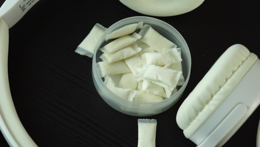 a top down view of an open round plastic container filled with small white nicotine pouches and snus pads on a dark wooden table surface surrounded by white overhead headphones