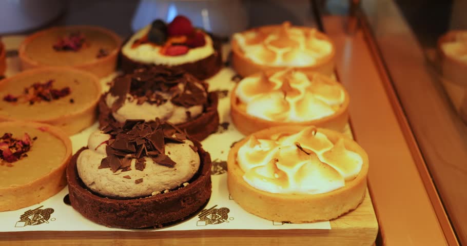 Assorted mini cakes put on tray in cafe dessert counter display. Sweet selection shows nut topping fruit slices with chocolate curls and soft cream swirls