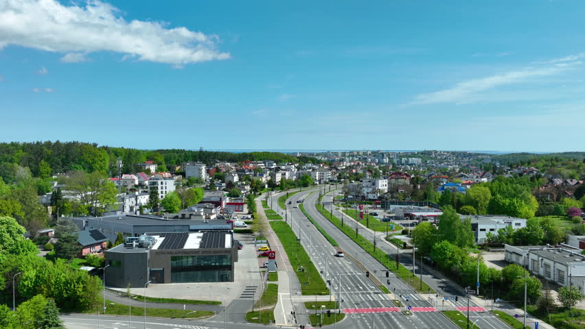 Cinematic aerial shot of Gdynia city suburbs featuring a main road with light traffic surrounded by lush green trees and residential buildings under a blue sky.