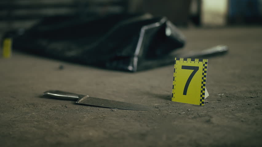 Close-up of knife and yellow evidence marker near body bag at crime scene