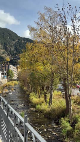 Mountain river flowing through Andorra landscape with autumn trees and peaceful urban nature scenery. Scenic stream surrounded by colorful foliage, mountain hills and small town architecture under soft daylight. Pyrenees travel destination, tranquil outdoor atmosphere and cinematic vertical footage.