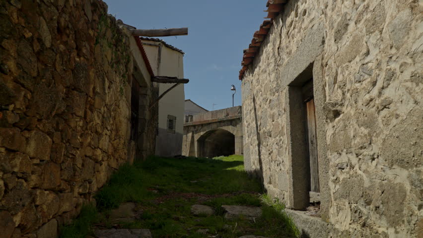 Rural footpath winding between ancient stone buildings, leading towards a historical arch bridge under a clear blue sky