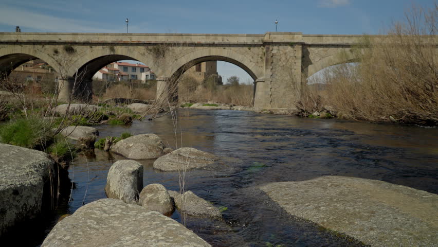 Ancient stone arch bridge spanning a river with rocks and natural vegetation, offering a view of rural buildings