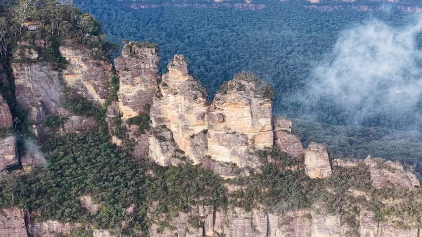 Close-up aerial shot of the Three Sisters rock formations in Katoomba, with dramatic clouds passing beside the cliffs, creating a cinematic and atmospheric mountain scene.