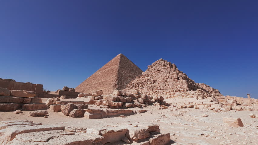 Handheld footage of the Queen Henutsen pyramid and the Great Pyramid of Giza surrounded by ancient stone ruins and scattered limestone blocks in the Egyptian desert. Giza plateau, Egypt