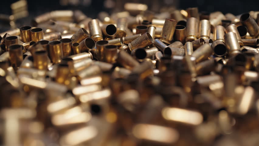 Macro close-up of a large pile of shiny brass spent bullet casings falling in slow motion, representing concepts of conflict, war, violence, and ammunition manufacturing