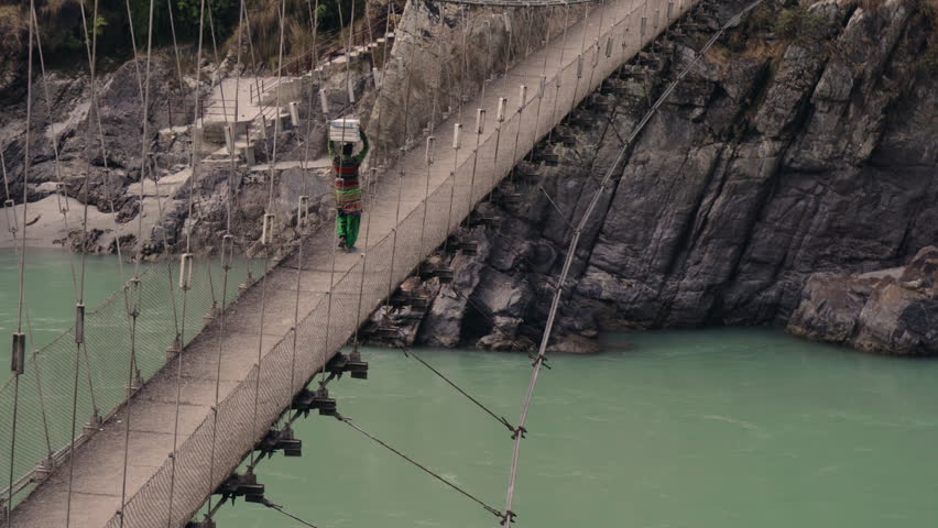 Aerial view of a Nepali woman walking across a precarious suspension bridge over a turquoise river while carrying a heavy metal box on her head, showcasing daily life in a rural village