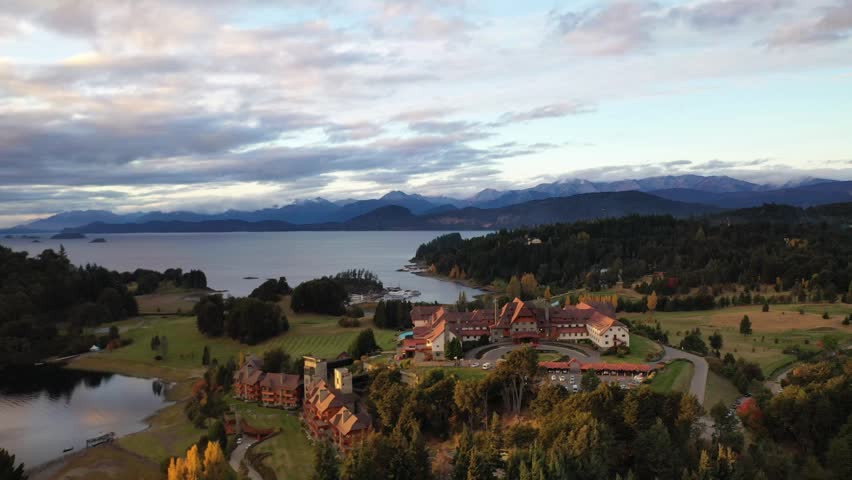 Breathtaking drone panorama of the Bariloche lake district at sunset. Showcases the deep blue water, forested islands, and rolling mountain ridges bathed in warm golden light under a cloudy sky.