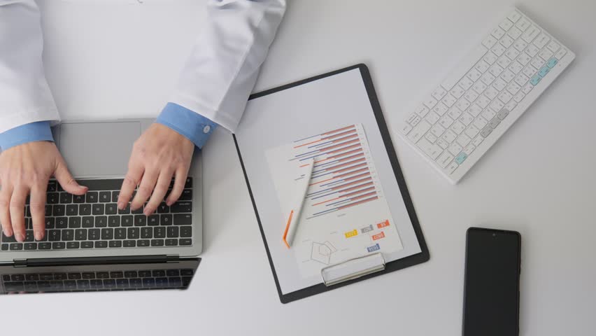 A physician types on a laptop surrounded by medical documents, a stethoscope, and analytical charts. Digital healthcare solutions and data driven decision making are highlighted in this workspace
