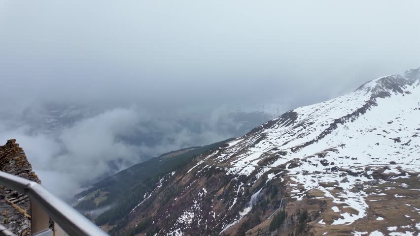 Mountain views from the Grindelwald First Cliff Walk as fog drifts through snowy alpine peaks and steep cliffs in the Swiss Alps above the valley below.
