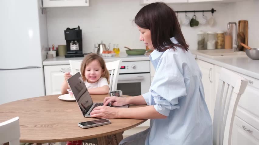 A mother works on her laptop at the kitchen table while her young daughter enjoys a snack. A cozy domestic scene showcasing work-life balance at home.