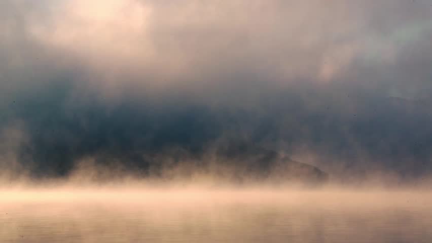 Vapor and clouds moving over a lake at sunrise, with a mountain hill peaking in the distance