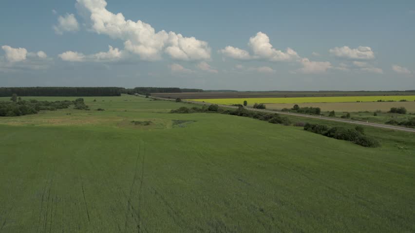 Wide aerial footage of a straight rural road cutting through green meadows and farmland under cumulus clouds, summer landscape in the Ural region, Russia