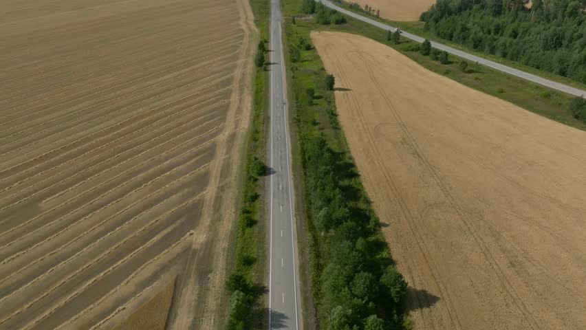 Top-down aerial footage of a straight asphalt country road running between golden harvested farmland and green trees in the Ural region, Russia, on a sunny summer day