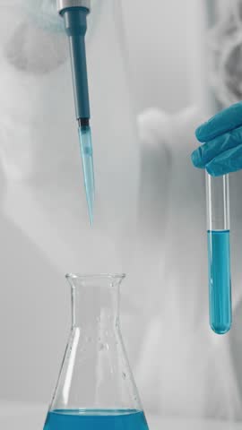 Researcher wearing full protective gear uses pipette to transfer blue solution into test tube, highlighting precision in laboratory experiments. Scene emphasizes scientific progress, safety