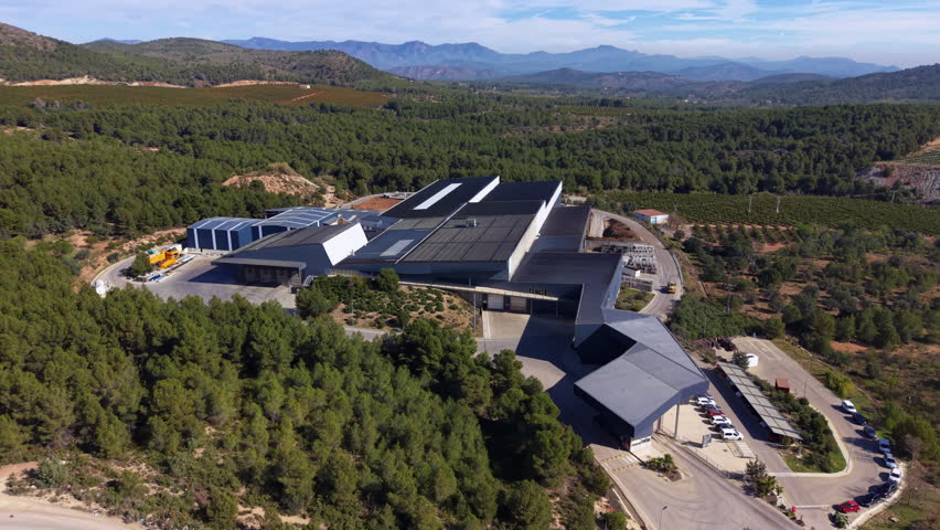 Aerial view of a modern waste processing and recycling facility nestled among pine forests and mountains in rural Spain, with solar panels on rooftop and industrial equipment