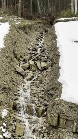 Small mountain stream melting and rushing down a rocky, eroded hillside covered with patches of white snow in early spring