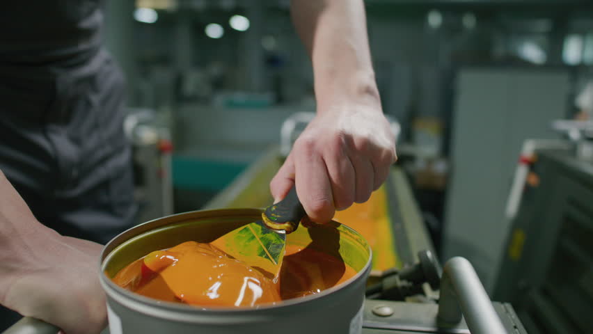 Hands of male industrial worker scooping vibrant yellow ink from bucket onto spatula knife in printing factory. Close up shot