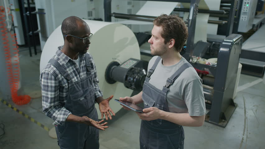 African American male technician explaining manufacturing process to Caucasian quality control inspector using digital tablet on production floor of printing plant. Tilt down shot
